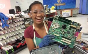 AJ-makerspace-KJ-CC3 | MiddleWeb Young woman wearing safety goggles and gloves smiles while holding a computer motherboard in a repair shop workshop.