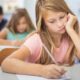 Young girl in a pink shirt writing at a desk in a classroom, with other students blurred in the background.